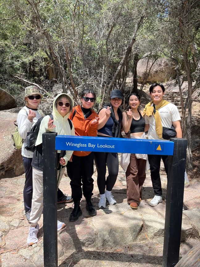 Family photo in front of the Wineglass Bay Lookout sign in Tasmania, in front of rocks in the bush.