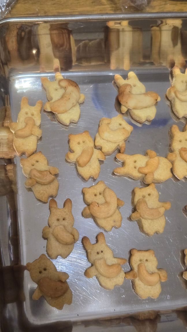 Cute cookies in the shape of a bear and a rabbit.  They're lying on a metal baking tray and each hugging a cashew nut.